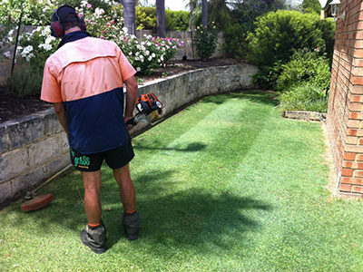 A Kiss My Grass crew member in hi-vis and ear protection whipper-snipping along a curved limestone garden edge