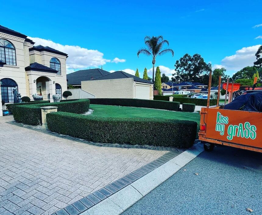 Squared-off hedges framing a mown lawn, with the orange Kiss My Grass trailer at the kerb